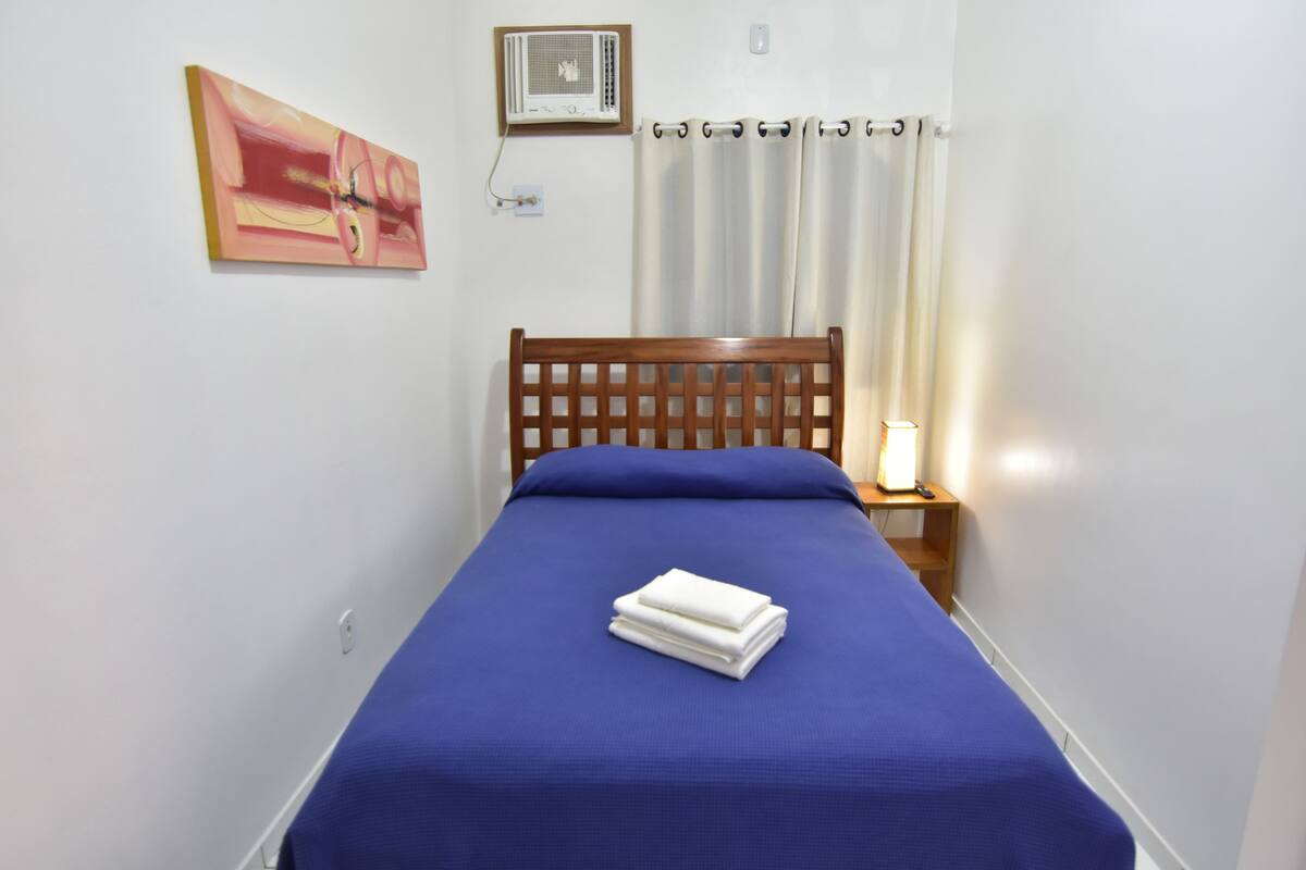 Una cama bien hecha con colcha azul y toallas blancas, cabecera de madera, lámpara de noche y aire acondicionado instalado en la pared.
