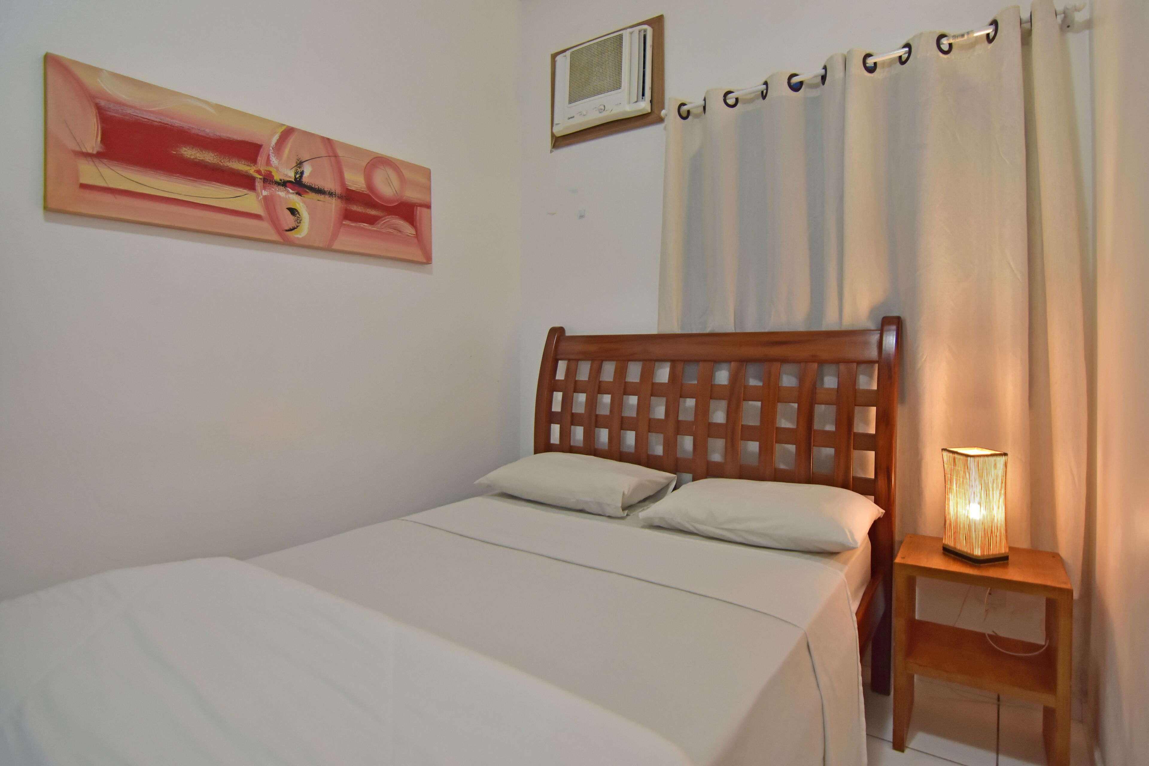 A bedroom with a wooden headboard, a bed with white bedding, a bedside lamp, a wall-mounted air conditioner, and a painting.