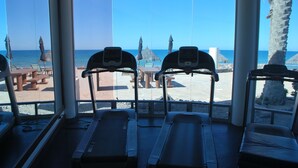 Academia com esteiras e equipamentos de ginástica, com vista para uma praia com palmeiras e guarda-sóis de palha.