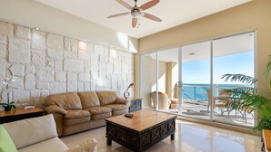 Uma sala de estar espaçosa com parede de pedra, sofá de couro marrom, mesa de centro de madeira e uma grande porta de vidro deslizante que dá para uma varanda com vista para o oceano.