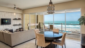 Un salón moderno con un televisor grande, un sofá, una mesa de comedor con sillas y vistas al océano a través de ventanas amplias.