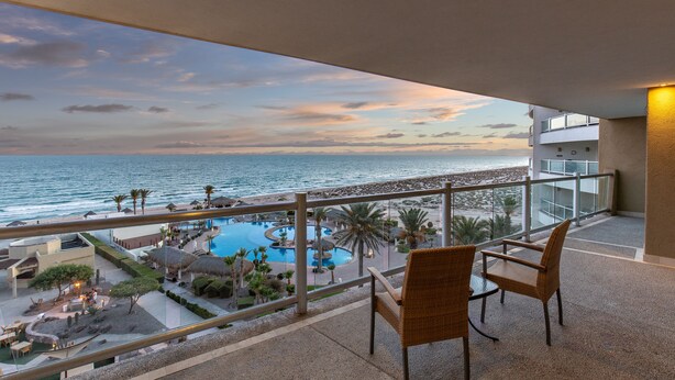 Balcón con sillas de mimbre, con vista a una piscina y zona de playa con palmeras.
