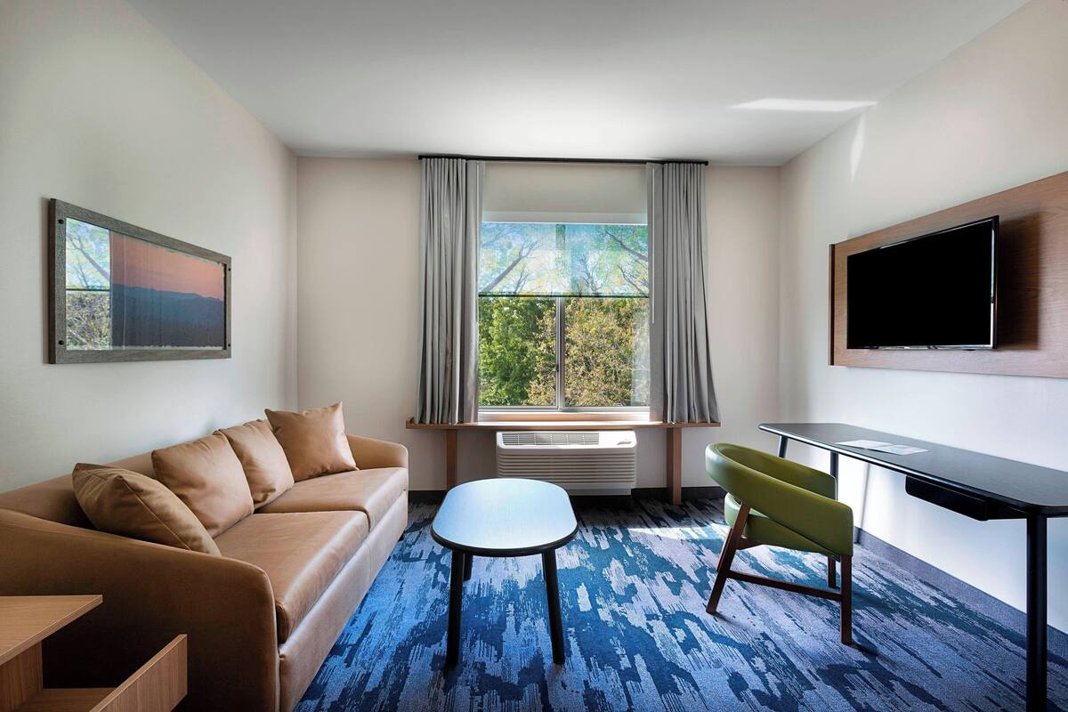 A modern hotel room with a sofa, a small table, a chair, a TV, and a window with curtains.