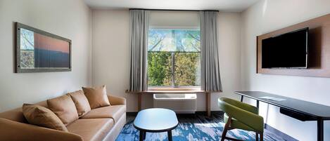 A modern hotel room with a sofa, a small table, a chair, a TV, and a window with curtains.