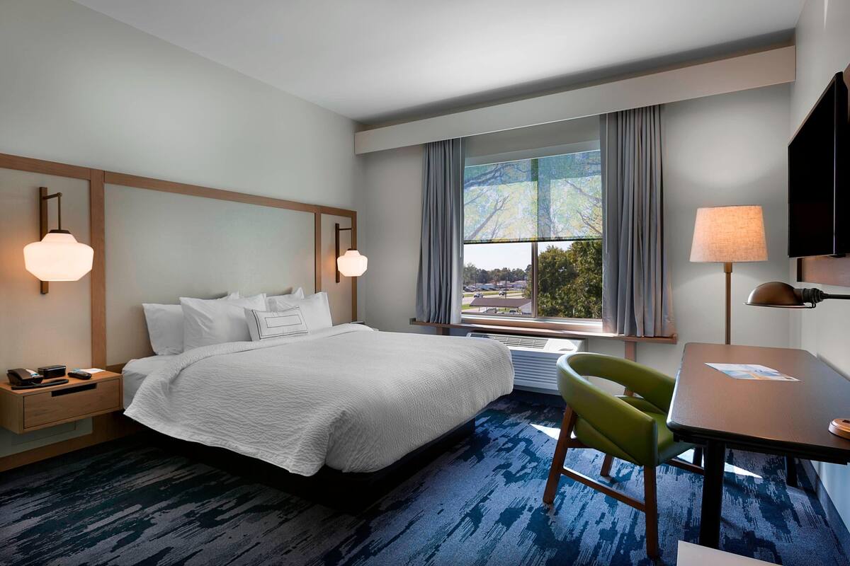 A hotel room with a large bed, a desk with a chair, a window with curtains, and a wall-mounted TV.