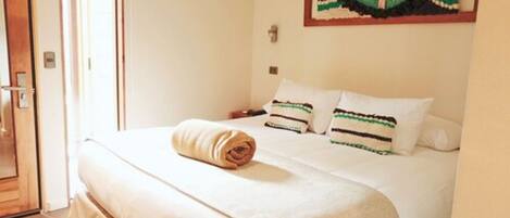 Una cama bien tendida con una toalla enrollada, una obra de arte en la pared y una puerta a la izquierda.