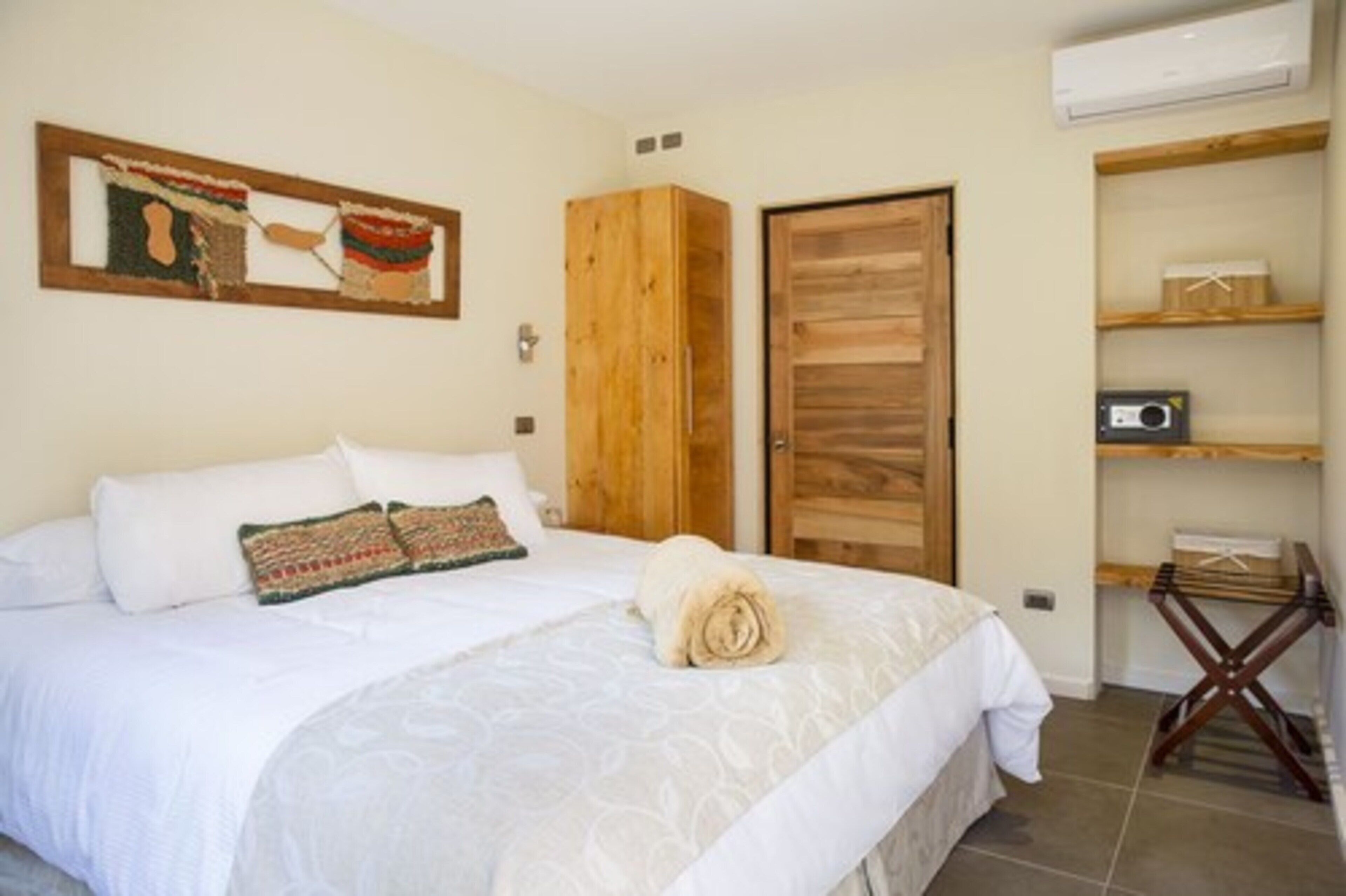 A hotel room with a bed, a wooden wardrobe, a wooden door, a shelf with a radio, and a wall-mounted air conditioner.