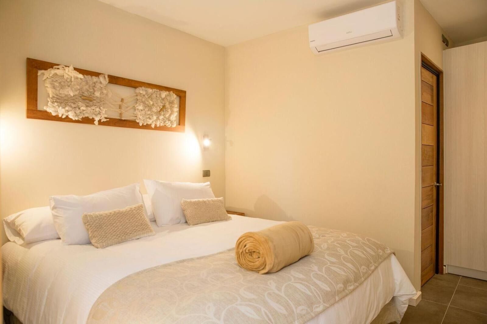 Une chambre d’hôtel avec un lit, des oreillers, une serviette roulée, un climatiseur fixé au mur et une porte en bois.