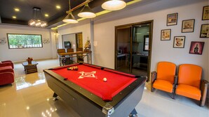 A pool table with a red felt surface, a billiards cue, and a white cue ball. There are two orange chairs and a red sofa in the room. - Baan Por Jai HuaHin Pool Villa (Hua Hin)