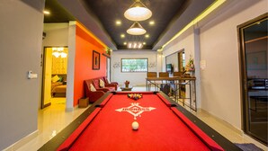 A pool table with a red felt, a chandelier, and a window with a view of greenery. - Baan Por Jai HuaHin Pool Villa (Hua Hin)