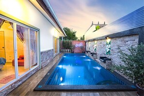 A swimming pool with a wooden deck, a brick wall, and a glass door leading to a room with a red chair. - Baan Jing Jai HuaHin Pool Villa (Hua Hin)