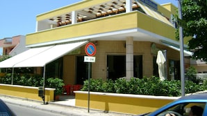 A two-story building with a yellow facade, a covered patio area, and a sign indicating a 24-hour cafe. - Oasi B&B (Porto Cesareo)