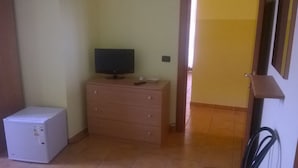 A small room with a white refrigerator, a wooden dresser with a flat-screen TV on top, and a door leading to another room. - Oasi B&B (Porto Cesareo)