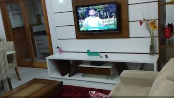 Uma sala de estar com uma televisão de tela plana presa a uma parede branca, um móvel branco para entretenimento, um sofá bege e um tapete vermelho.