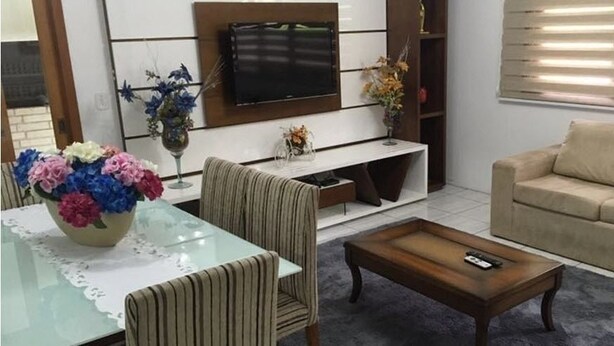 Un salon comprenant un canapé, une table basse et un téléviseur fixé au mur. On y trouve également une table à manger avec des chaises et un vase de fleurs.