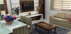 Uma sala de estar com sofá, mesa de centro e televisão na parede. Há uma mesa de jantar com cadeiras e um vaso de flores.
