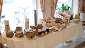A breakfast buffet with various food items and beverages on a table, set in a room with large windows and curtains. - Hotel Villa Bernt (Grado)