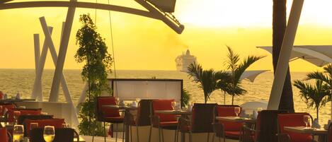 An outdoor dining area with tables and chairs, overlooking the sea and a cruise ship.