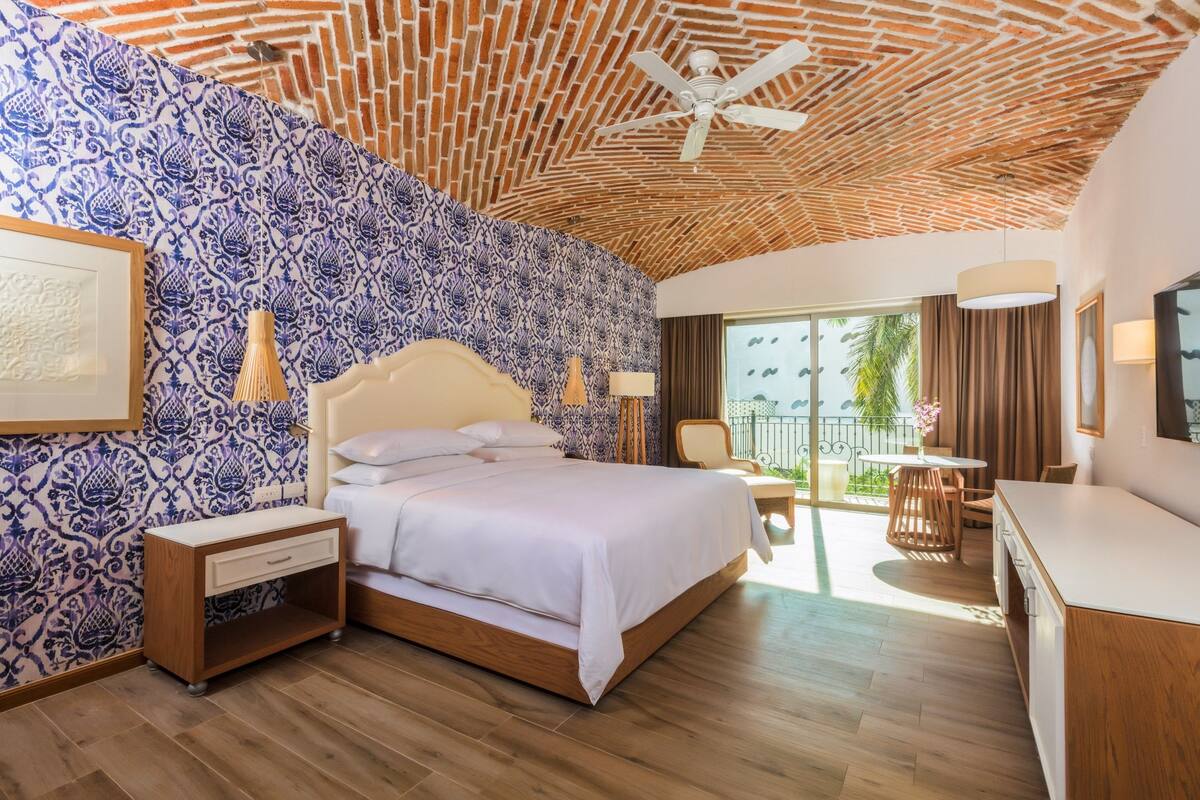 Une chambre avec un mur décoré de motifs, un lit, une table de chevet, un bureau et un balcon avec vue.