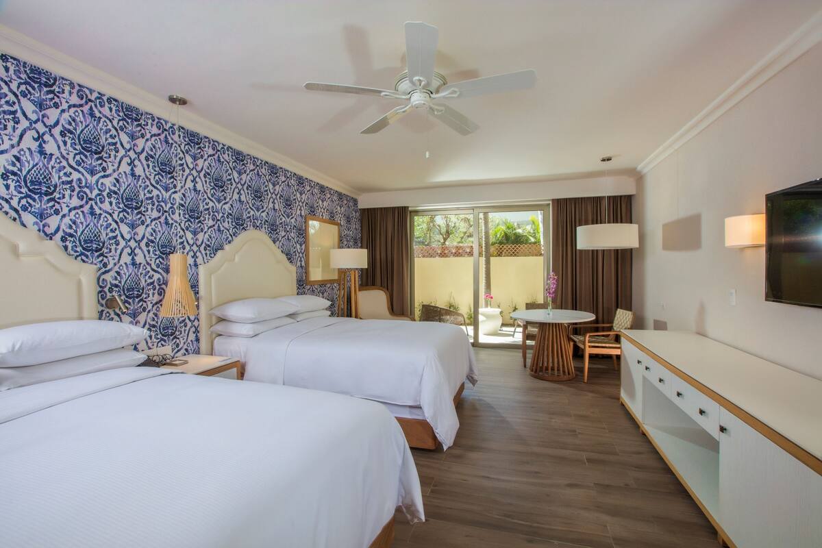 Une chambre d’hôtel avec deux lits, un ventilateur de plafond, une télévision, une petite table avec des chaises et une vue sur l’extérieur.