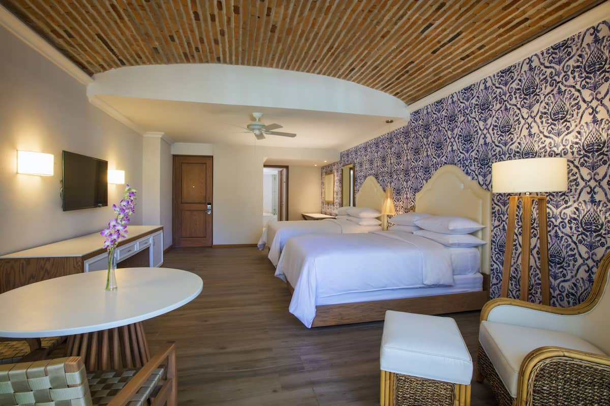 Une chambre d’hôtel avec un plafond en bois, un grand lit avec du linge de lit blanc, une petite table ronde, un canapé et un mur recouvert d’un papier peint à motifs bleus et blancs.