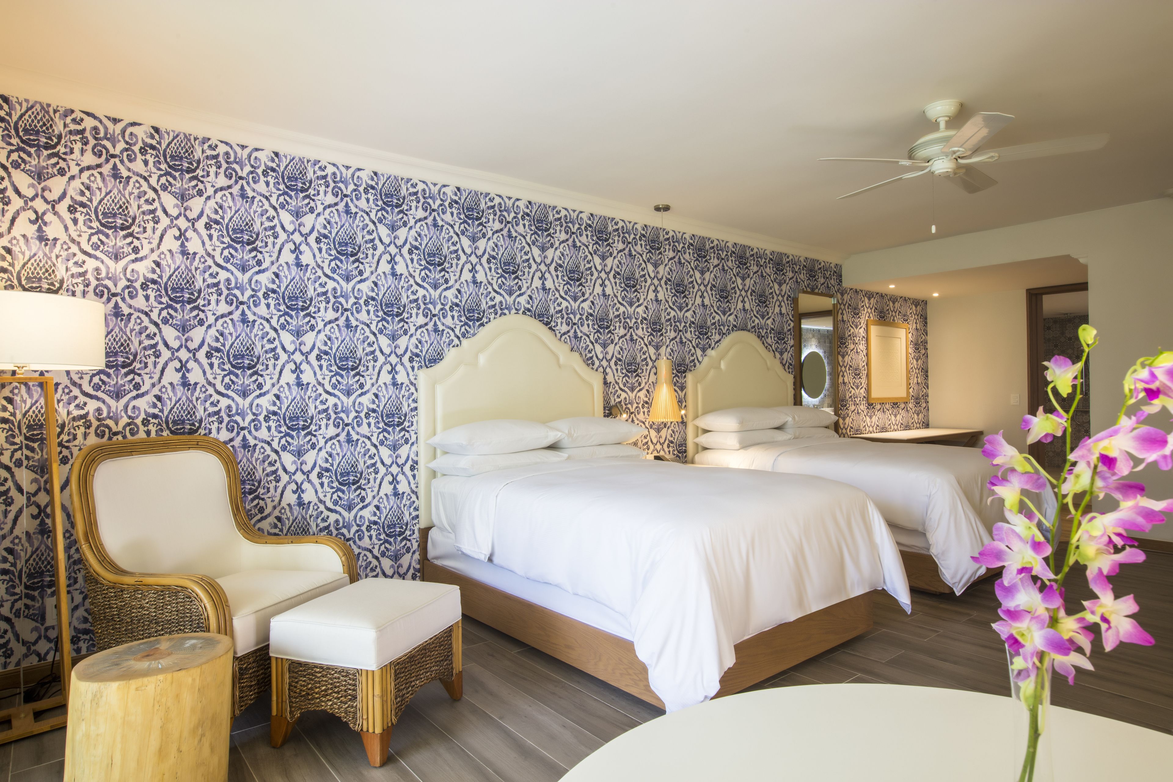 A hotel room with a patterned wallpaper, a bed with white bedding, a chair, a small table, and a vase with flowers.