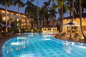 Una piscina con sillas de descanso y sombrillas, rodeada de palmeras y edificios de hotel.