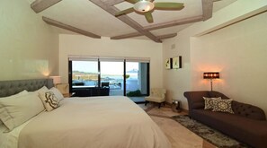 A bedroom with a large bed, a sofa, a ceiling fan, and a view of the outdoors.