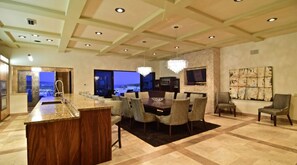 A spacious living area with a large dining table, comfortable seating, a kitchen area, and a view of the cityscape.