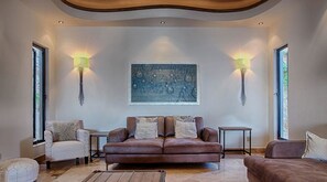 A living room with a brown leather sofa, two white armchairs, and a coffee table. There are two wall-mounted lamps and a large abstract painting above the sofa. - Villa Vegas by Cabo Platinum (Cabo San Lucas)