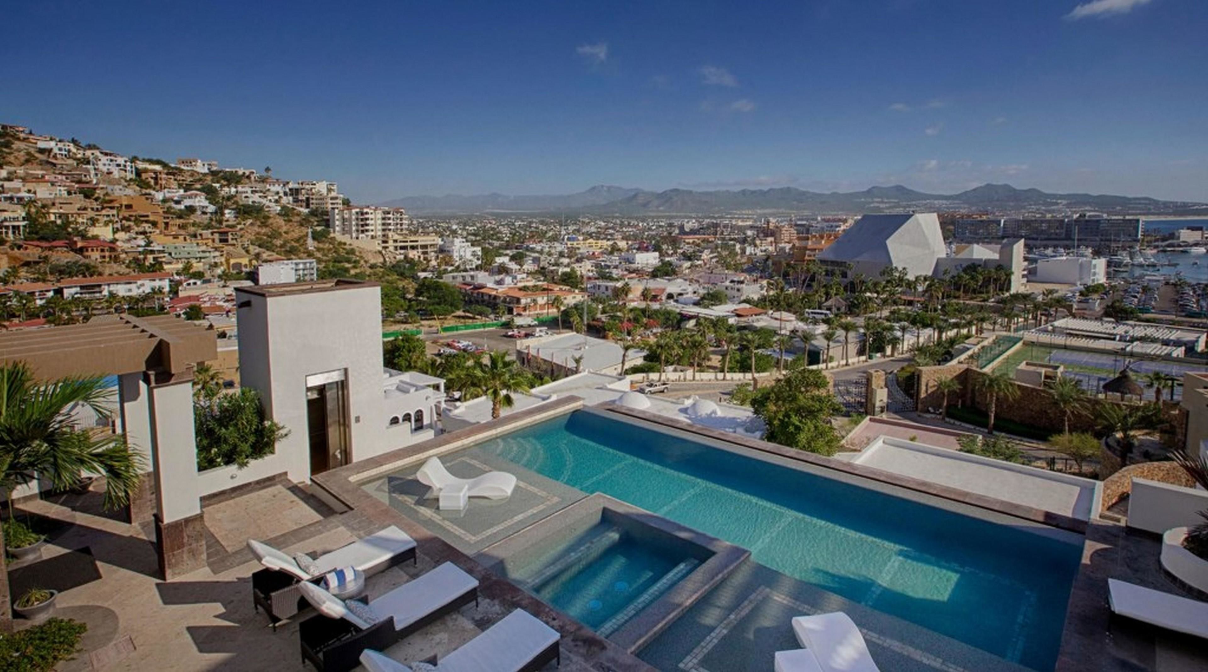 A rooftop pool with lounge chairs, a view of a coastal cityscape, and a mountainous backdrop.