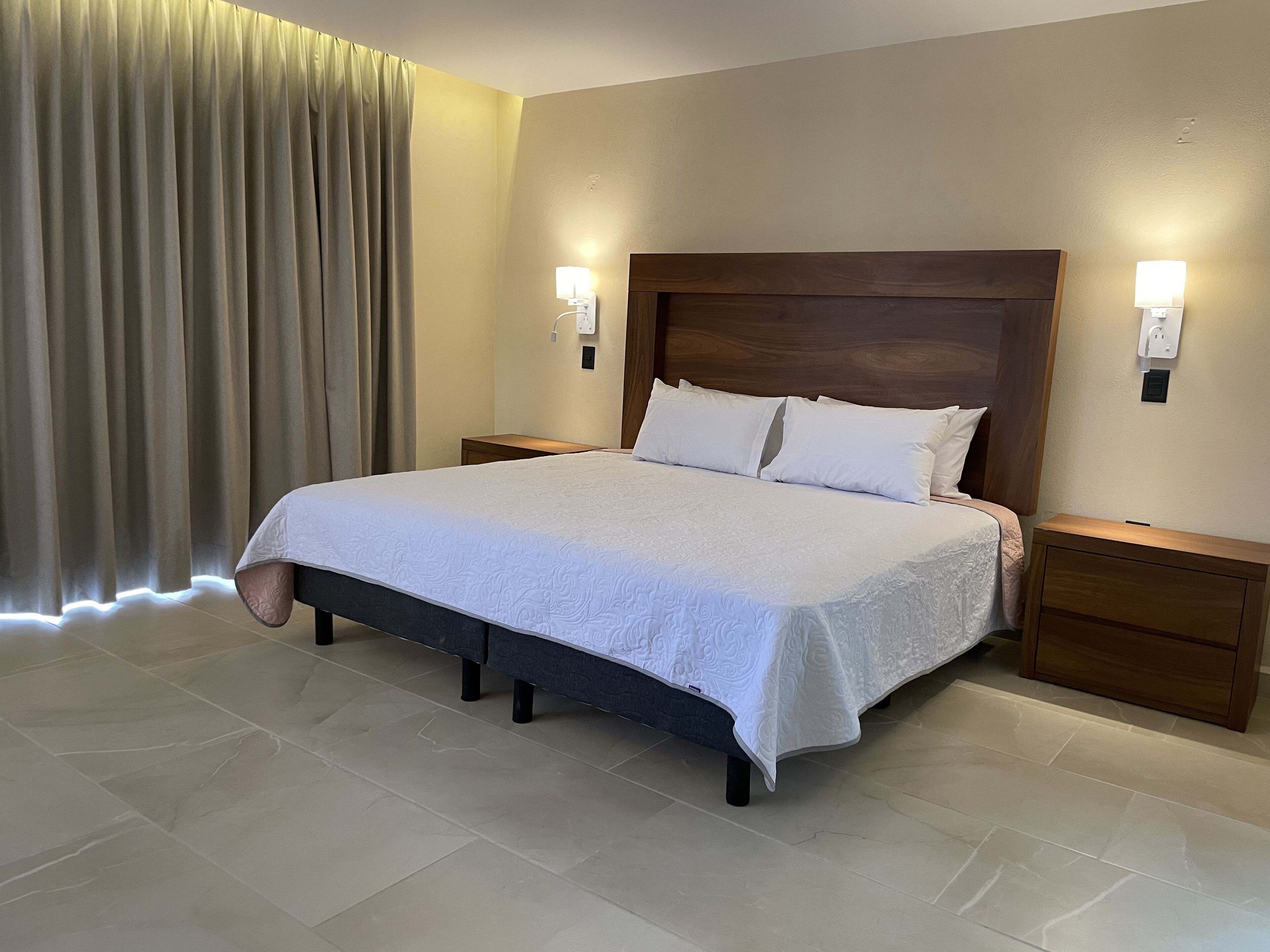 A neatly made bed with a white bedspread and two pillows, flanked by wooden nightstands with wall-mounted lamps.