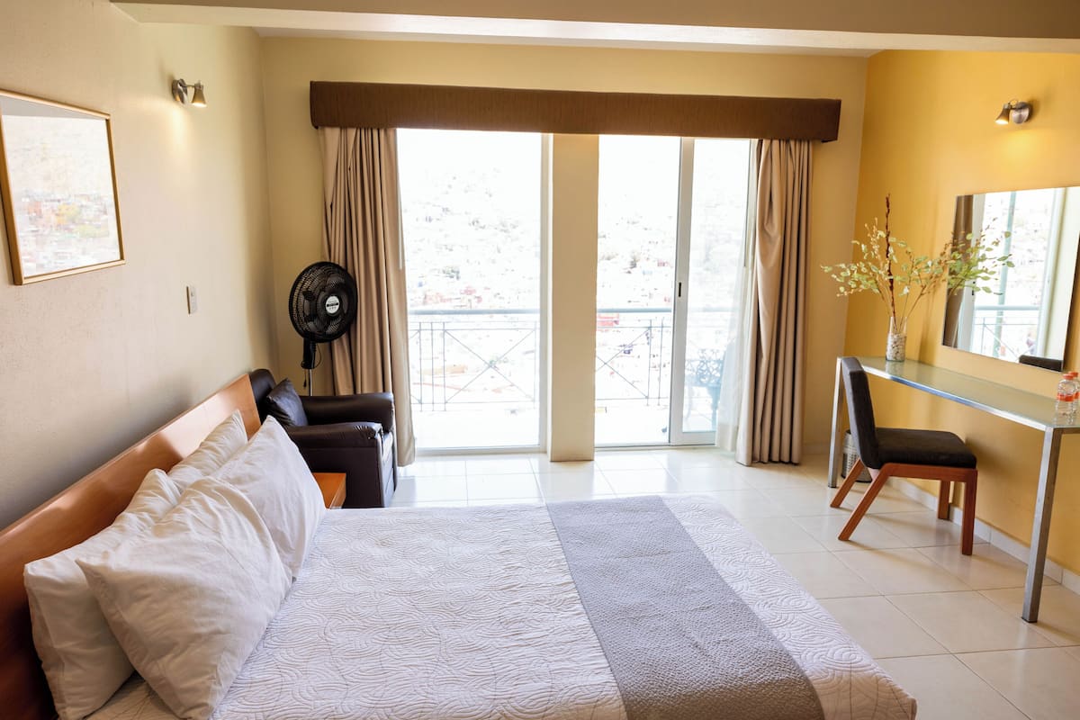 A hotel room with a bed, a desk, a chair, a fan, and a view of the outside through large windows.