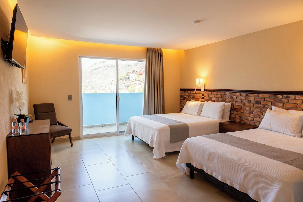 A hotel room with two beds, a desk, a chair, and a balcony with a view of the mountains.