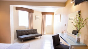 A hotel room with a bed, a sofa, a chair, a glass table, a mirror, and a window with curtains.