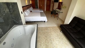 A hotel room with a large bed, a separate bed, a desk, a chair, a black sofa, a bathtub, and a mirror.