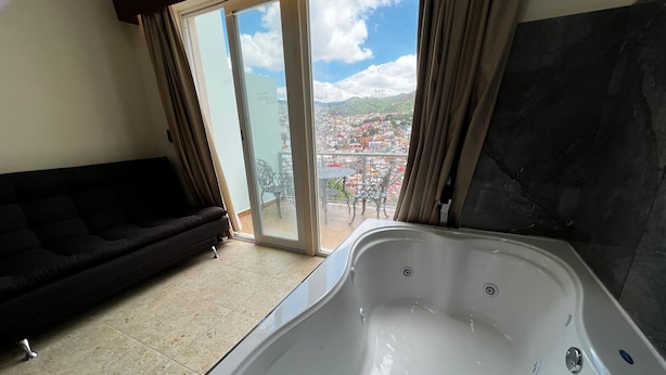 A room with a black sofa, a large bathtub, and a sliding glass door leading to a balcony with a city view.