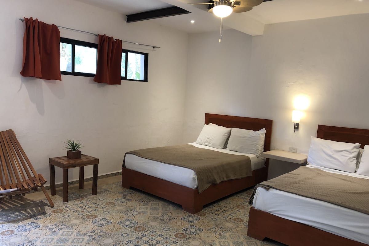 Un dormitorio con dos camas, un ventilador de techo, una mesita con una planta y una ventana con cortinas.