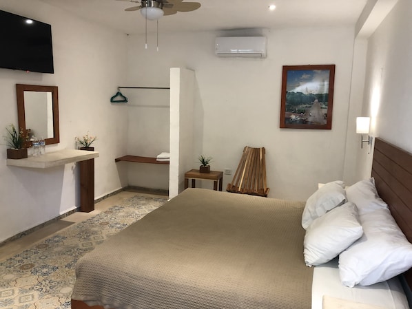Una habitación de hotel con cama, mesita de noche, espejo, televisor, aire acondicionado y un cuadro en la pared.