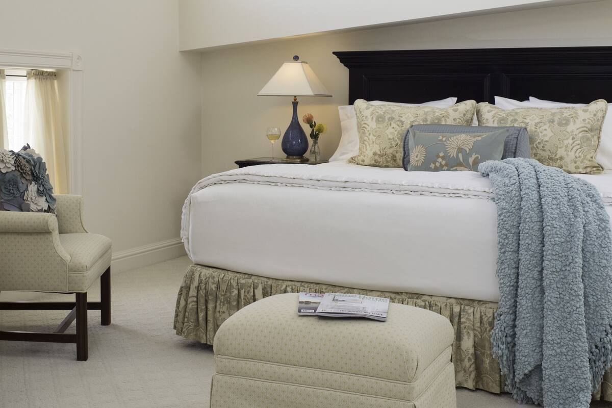 A neatly arranged bedroom with a bed, a chair, a nightstand with a lamp, and a window with curtains.