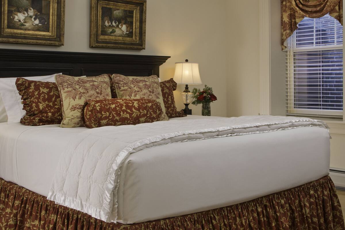 A neatly made bed with a white duvet and brown floral-patterned pillows, a dark wooden headboard, and a vase with red flowers on a bedside table.
