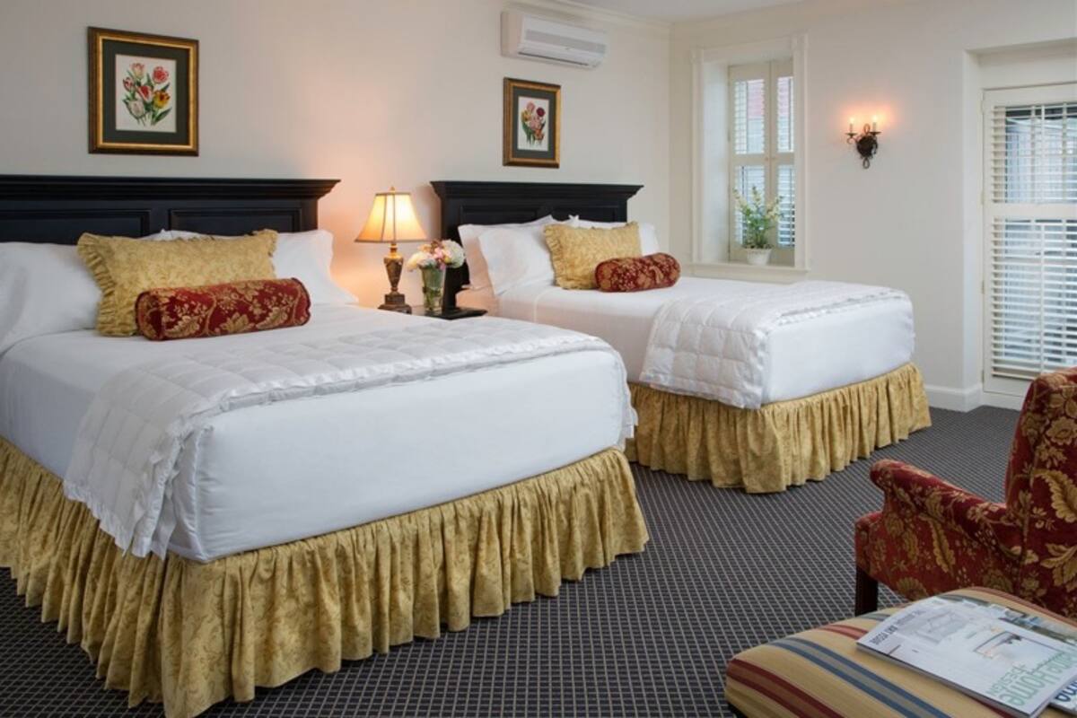 A hotel room with two beds, each with a white bedspread and gold trim. There are two wall-mounted lamps, a window with blinds, and a chair with a floral pattern.