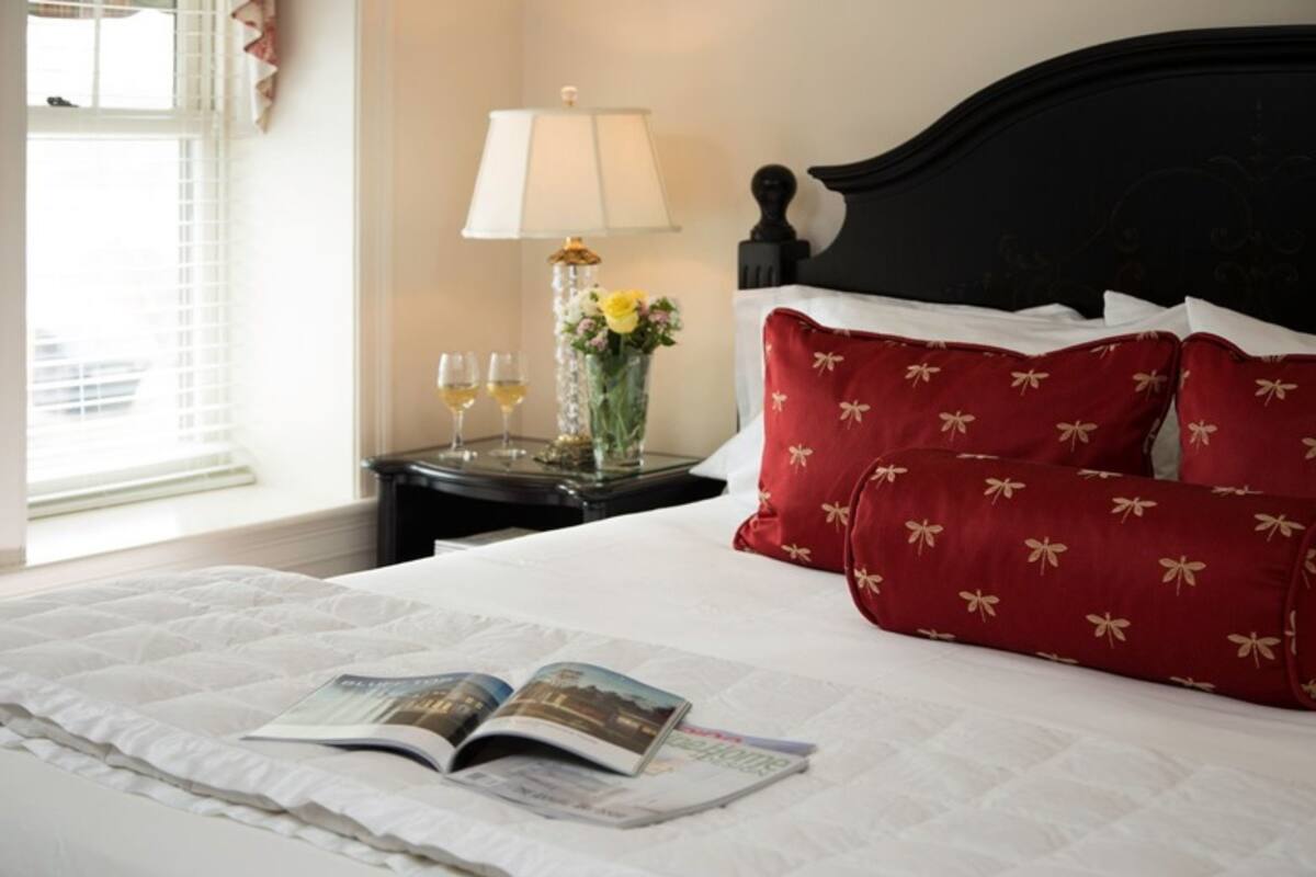 A neatly made bed with a magazine, a vase with flowers, and two glasses of wine on a nightstand.