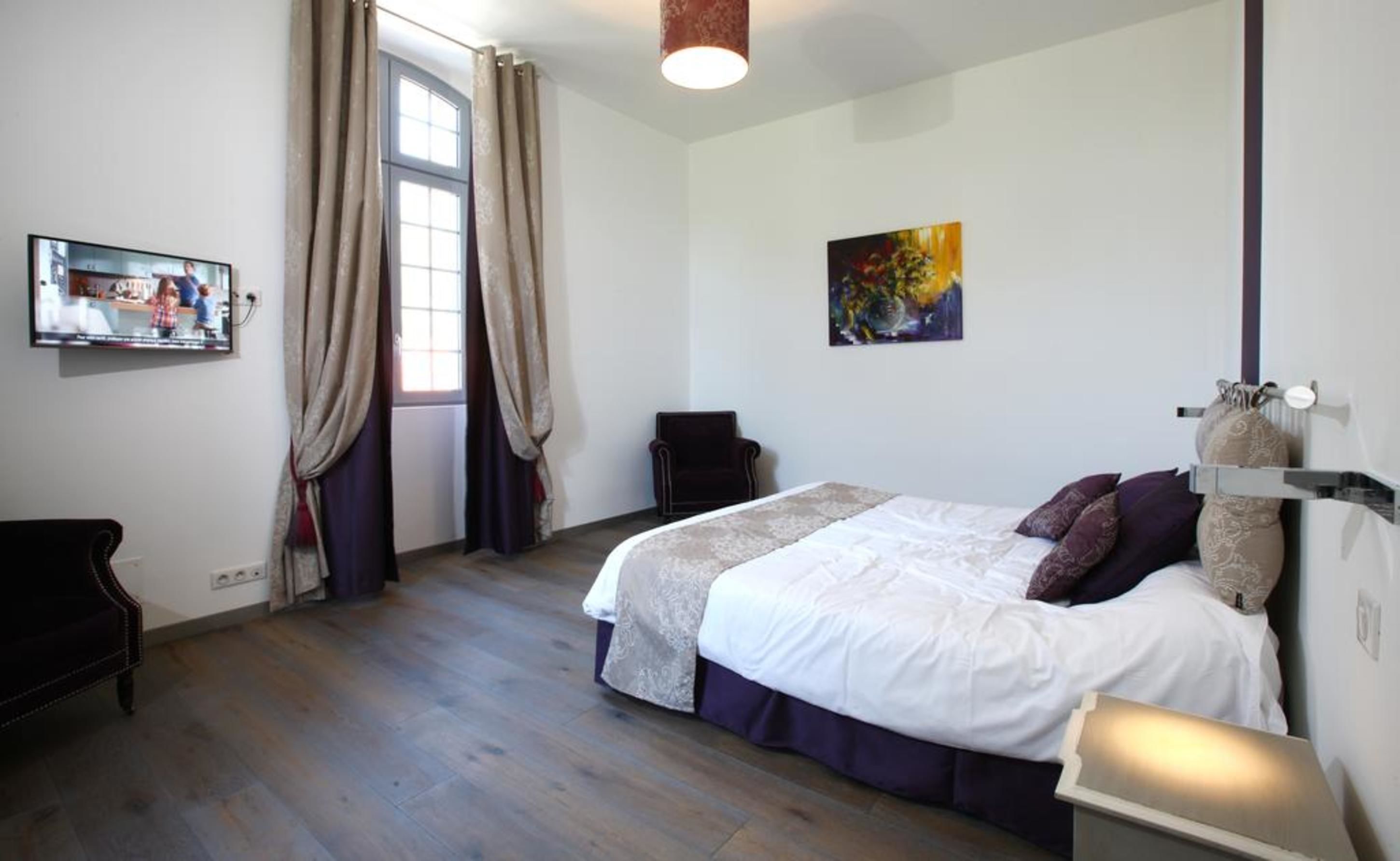 A bedroom with a bed, a chair, a painting, and a television on the wall.