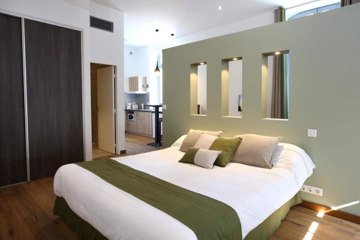 Une chambre à coucher moderne avec un lit, une literie verte et blanche, une cuisine en arrière-plan et un grand miroir sur le mur.