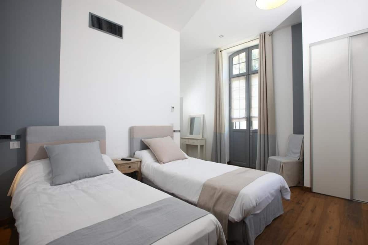 Une chambre d’hôtel avec deux lits simples, un bureau, une chaise, une armoire et une fenêtre avec des rideaux.