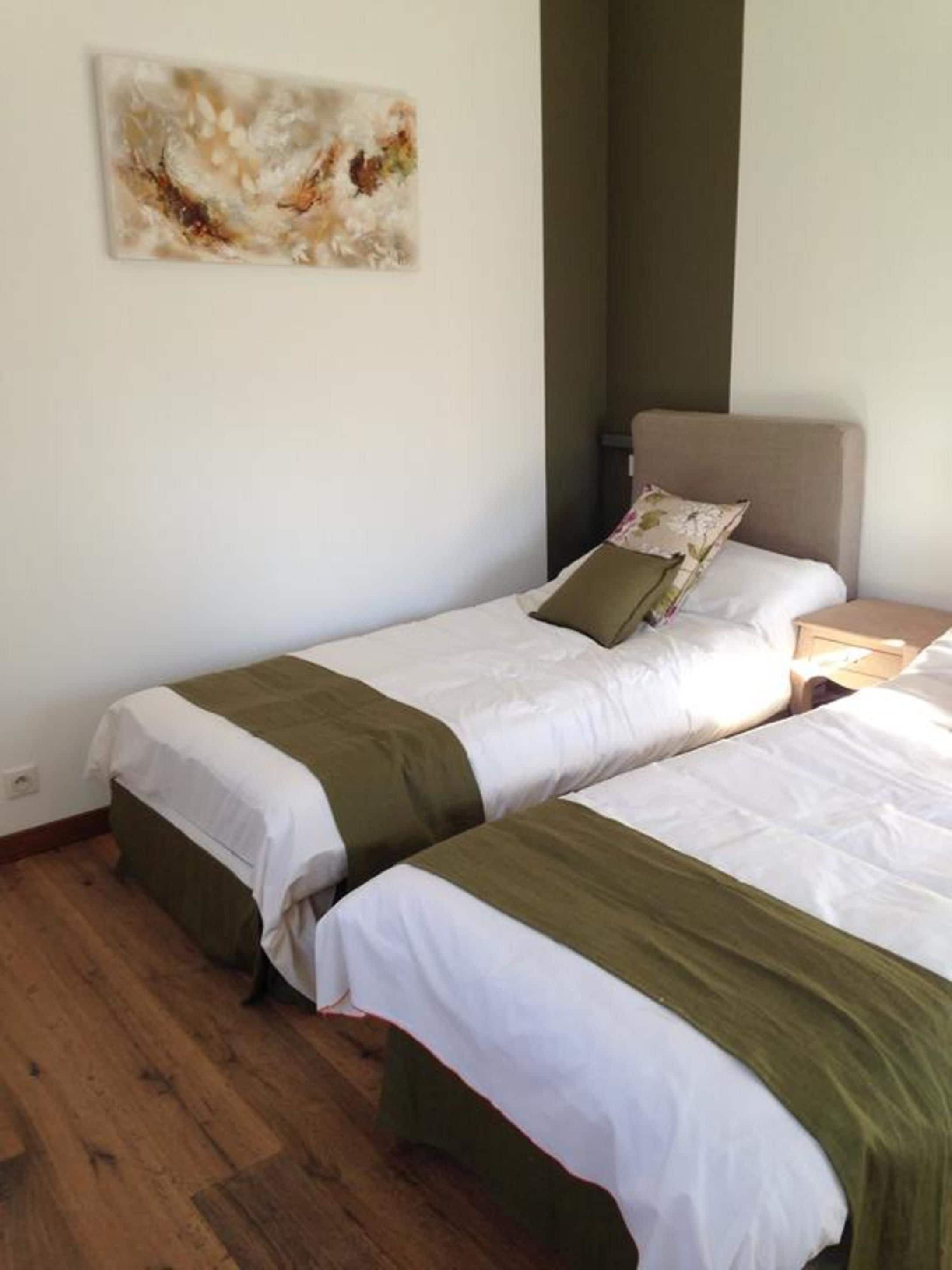 Una habitación con dos camas, cada una con ropa blanca y detalles verdes, suelo de madera, una mesita de noche y un cuadro en la pared.