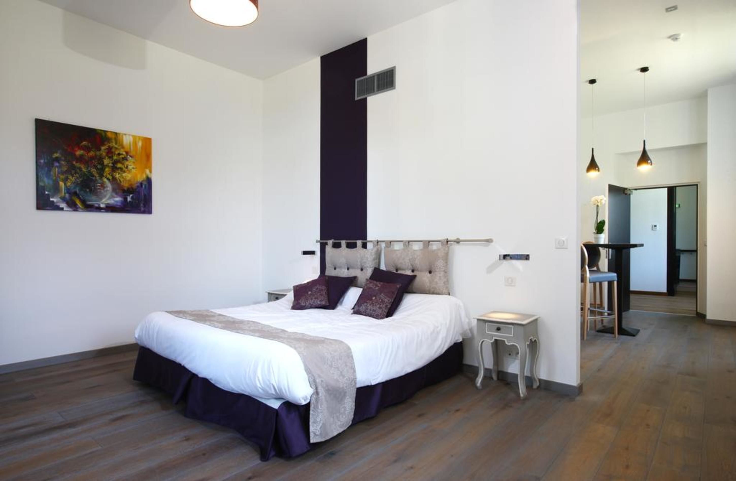 A modern bedroom with a large bed, a small bedside table, a chair, and a painting on the wall.