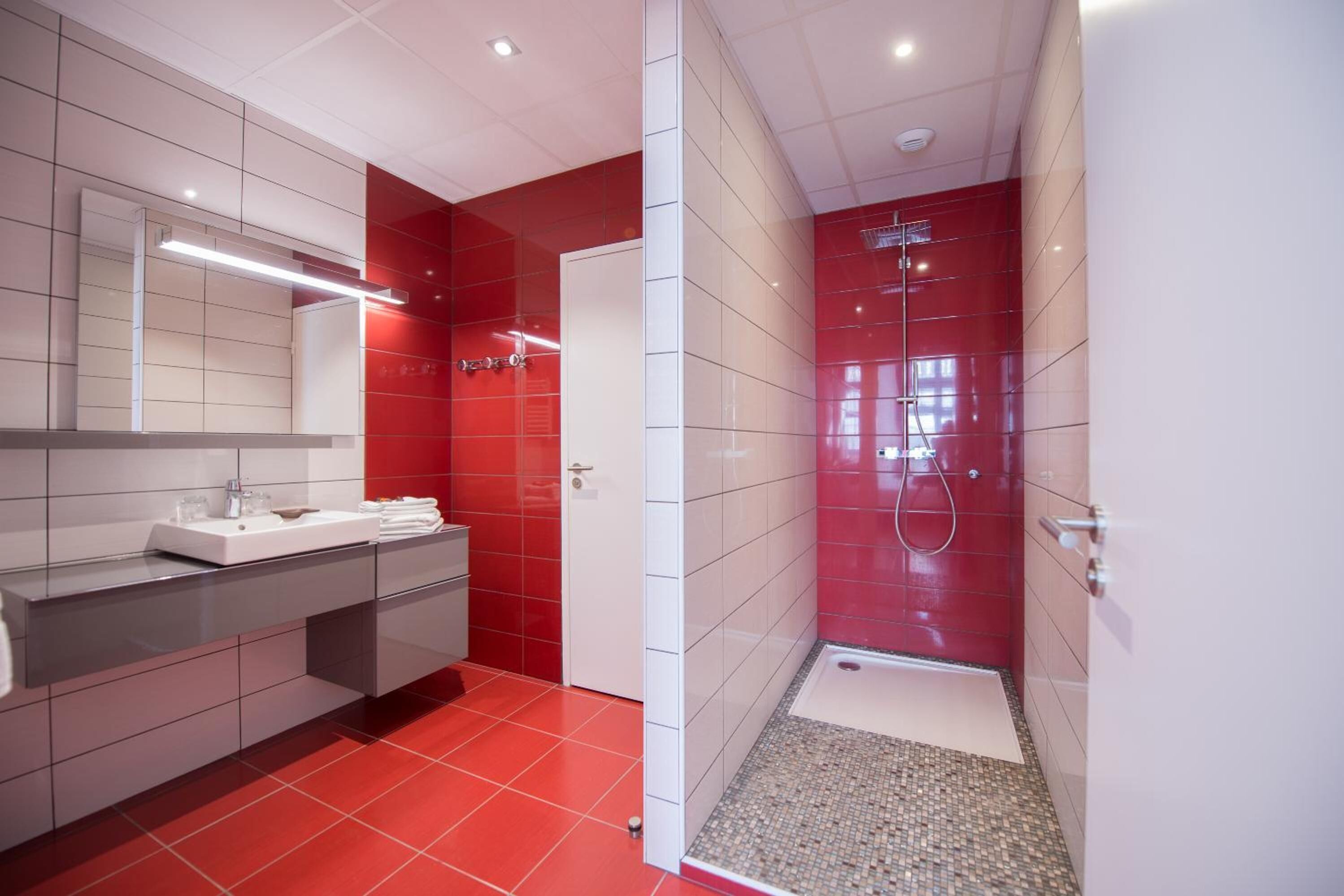 Un baño moderno con una decoración en tonos rojo y blanco, que incluye un lavamanos, espejo y zona de ducha.