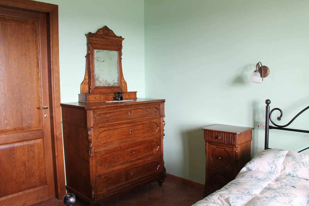 Une pièce comprenant une commode en bois, un lit avec une literie à motifs floraux, une petite table de chevet et un luminaire fixé au mur.